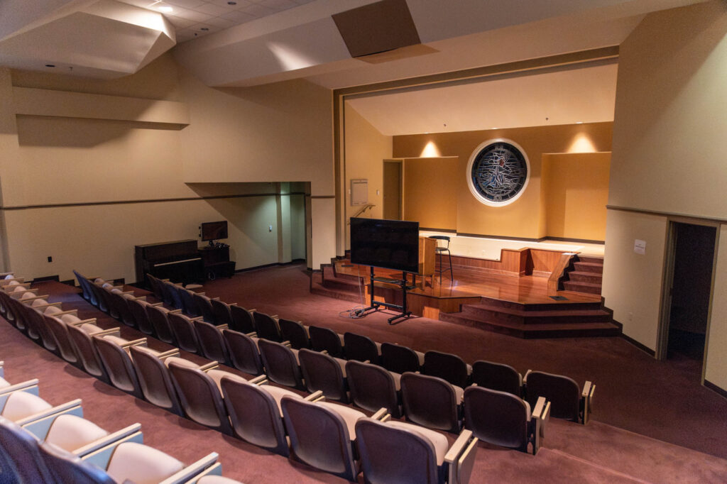 An empty auditorium features rows of beige seats, a small raised stage with a black monitor, a piano to the left, and a large circular decorative element on the back wall under soft lighting.