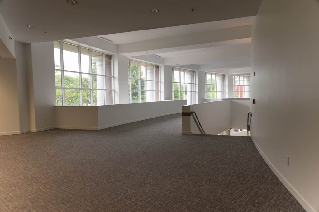 A spacious, carpeted hallway with large windows along one wall letting in natural light. A staircase with metal handrails leads down to a lower level. The walls and ceiling are painted white.