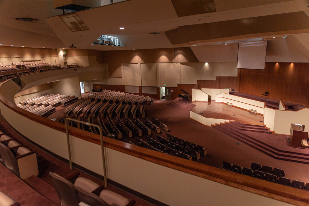 Large, empty auditorium with rows of beige seats, a wooden stage area, brown carpet, and multiple entrances. Overhead lighting illuminates the space, and there is a lectern on the stage. The view is from the upper level.