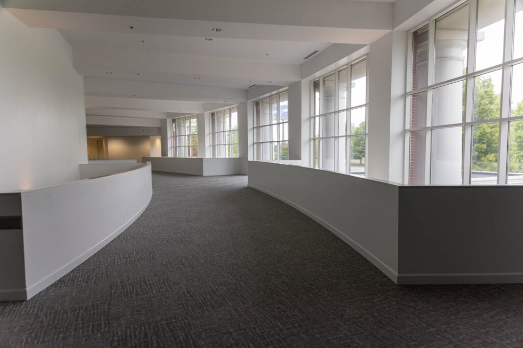 A wide, carpeted hallway with curved white walls and large windows on the right side, letting in natural light. The space appears clean and modern, with trees visible outside through the windows.