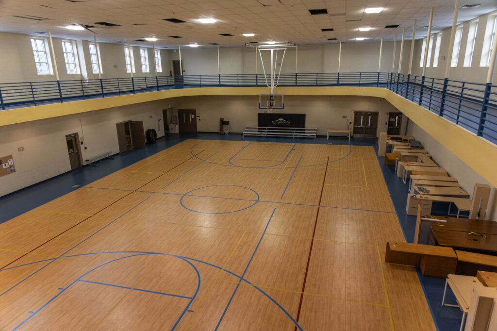 An empty indoor gymnasium with a basketball court, blue and yellow walls, upper-level track, several tables and chairs along the side, and fluorescent lighting overhead.