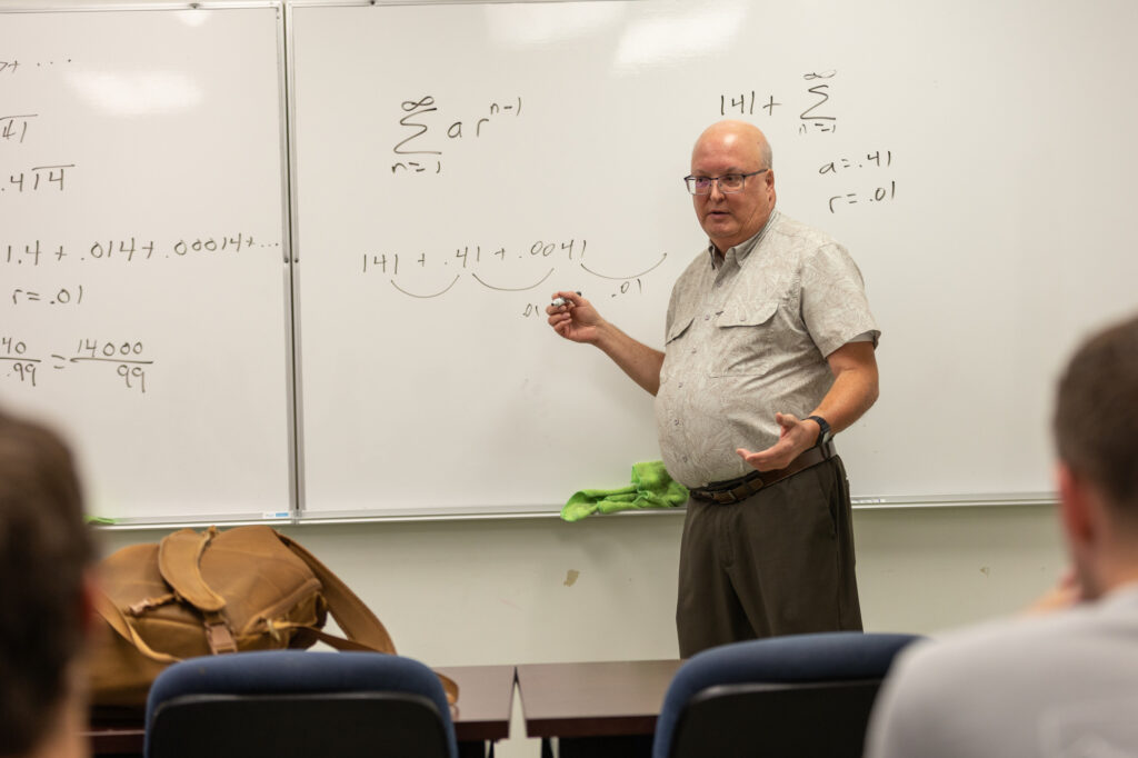 A man stands at a whiteboard covered with mathematical equations, holding a marker and gesturing as he teaches. Several students sit facing him, listening and observing the lesson. A brown bag is on the table.