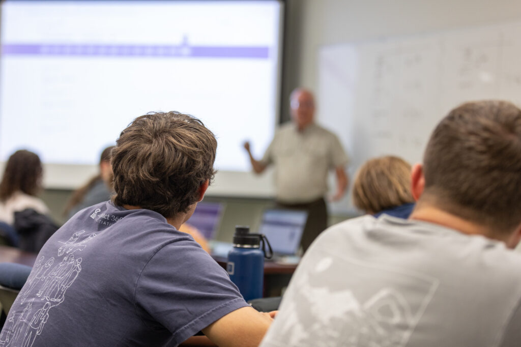 Students sit at desks in a classroom facing a teacher who is standing and speaking in front of a projection screen and whiteboard. Some students have laptops and water bottles on their desks.