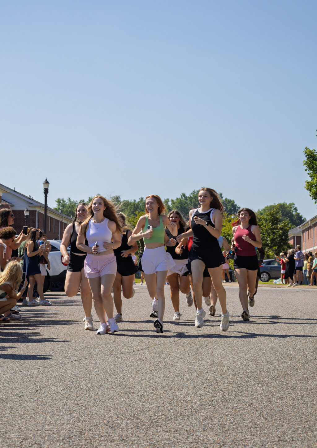 A group of young women are running on a paved street outdoors, surrounded by people watching on both sides. Buildings and trees are visible in the background under a clear blue sky.