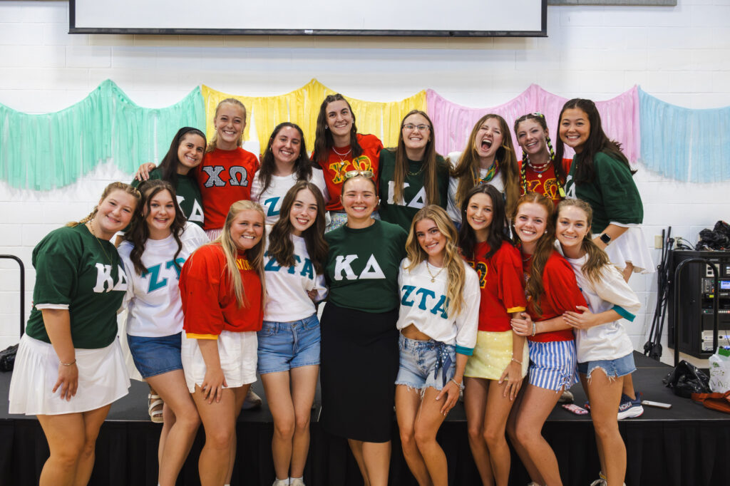 A group of sixteen young women wearing shirts with Greek letters pose together indoors in front of pastel streamers, smiling at the camera. Some women stand while others kneel or crouch in front.