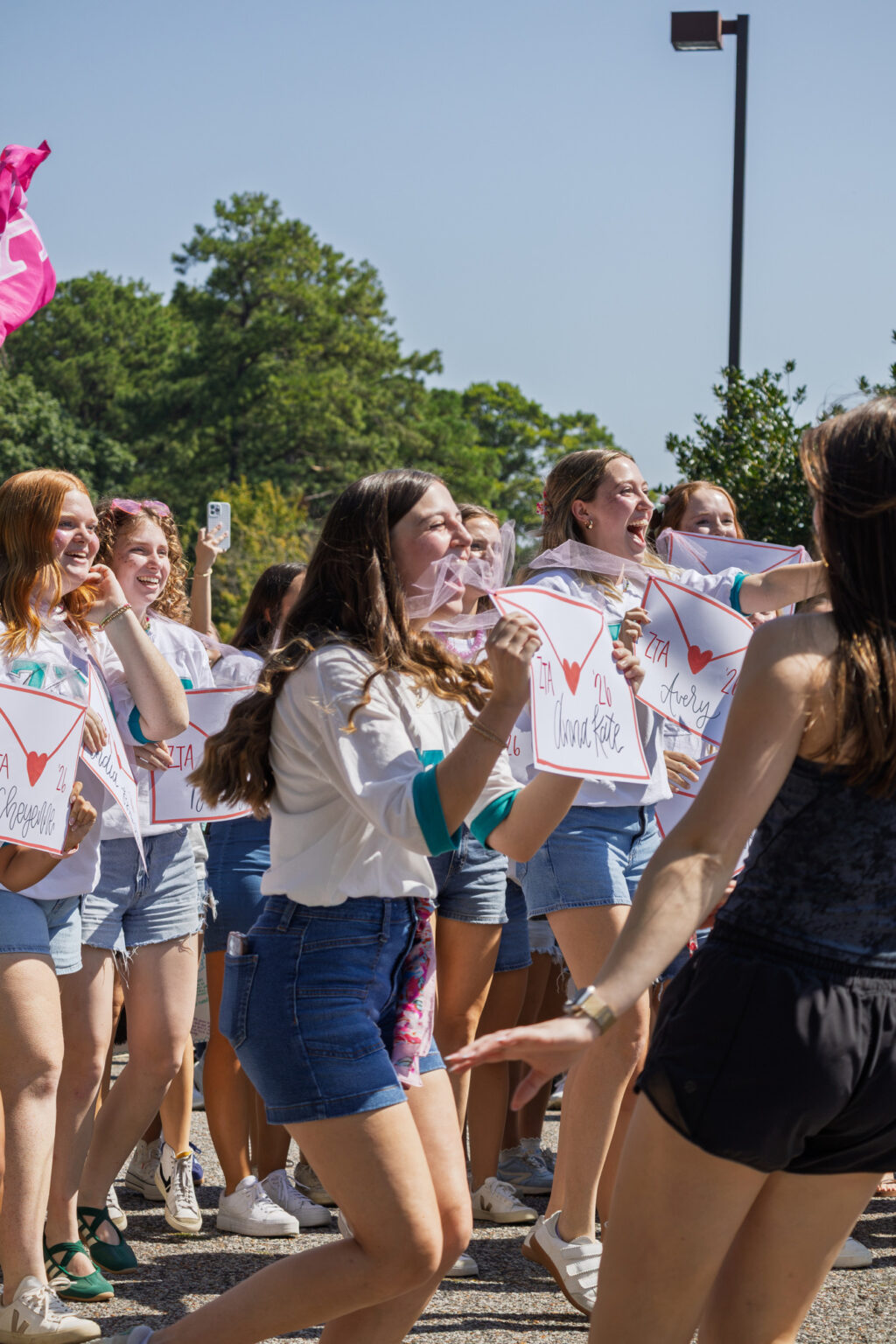 A group of young women outdoors, holding signs and smiling. They are wearing casual clothes, including denim shorts and white tops. Trees and a lamp post are visible in the background. It is a sunny day.