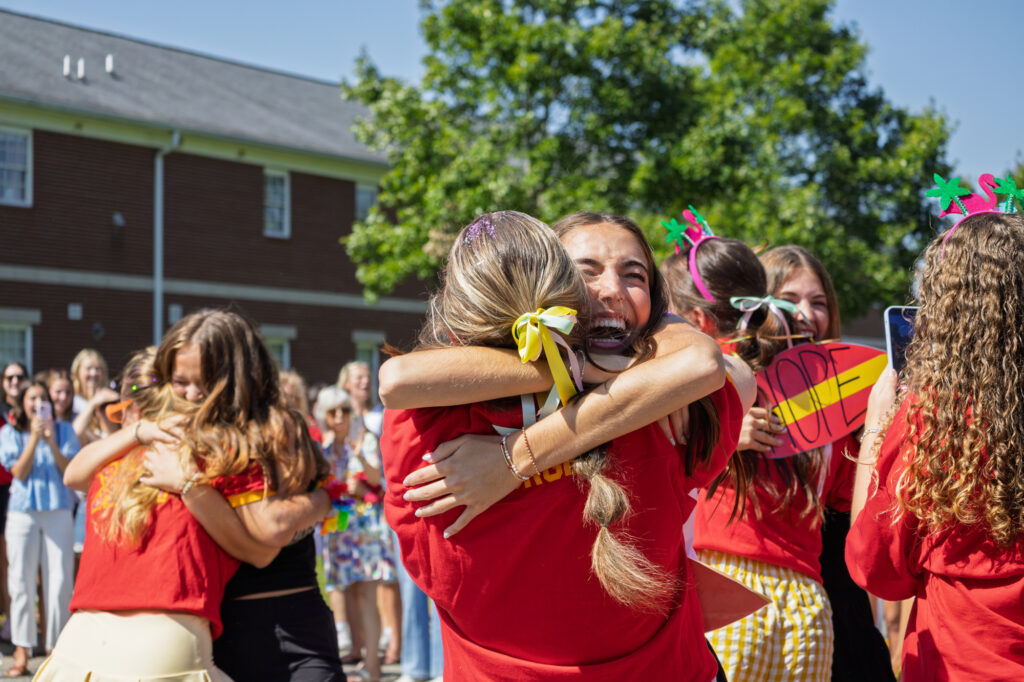 A group of young women outdoors hug and smile, wearing matching red shirts and hair ribbons. A crowd watches in the background. Trees and a brick building are visible under a clear sky.