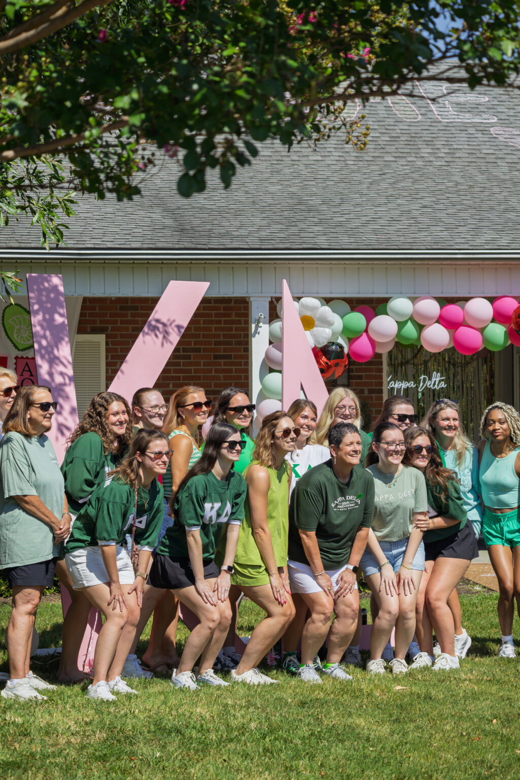 A group of women pose outdoors in front of large pink letters and a house decorated with balloons. Most wear green shirts and shorts. It is sunny and they are smiling for the photo. Some trees are visible at the top of the image.