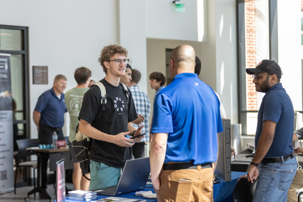 A young man with glasses stands at a booth, speaking to three men. Laptops, brochures, and a blue tablecloth are on the table. Other people and booths are visible in the background of a bright, sunlit room.