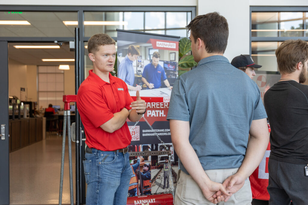 A man in a red polo shirt talks to another man in a gray shirt at an indoor event. They are standing near a career path poster, with other people and a glass wall visible in the background.