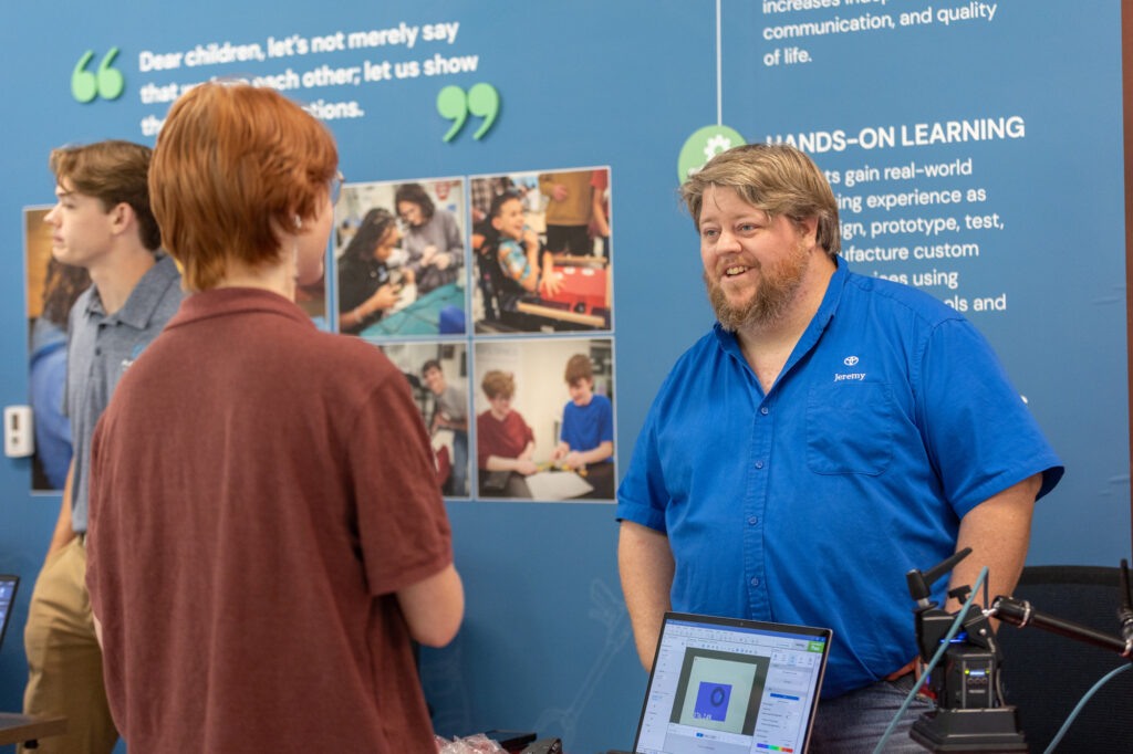 A man in a blue shirt talks to a red-haired person at a booth with technology equipment. A wall behind them displays educational quotes, photos, and information about hands-on learning.