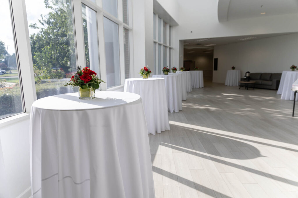 A bright room with tall windows, several round cocktail tables covered with white tablecloths, and small floral centerpieces. Some couches are seen in the background, and sunlight casts shadows on the light-colored floor.