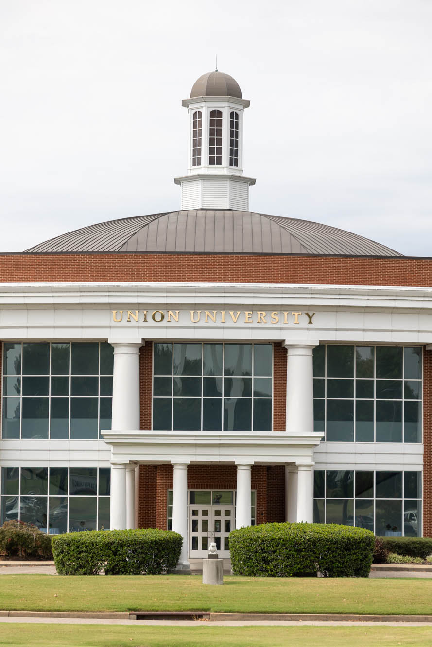 A large brick building with white columns, a central dome, and Union University written in gold letters above the entrance. Well-maintained shrubs and grass are in front of the windows and doors.