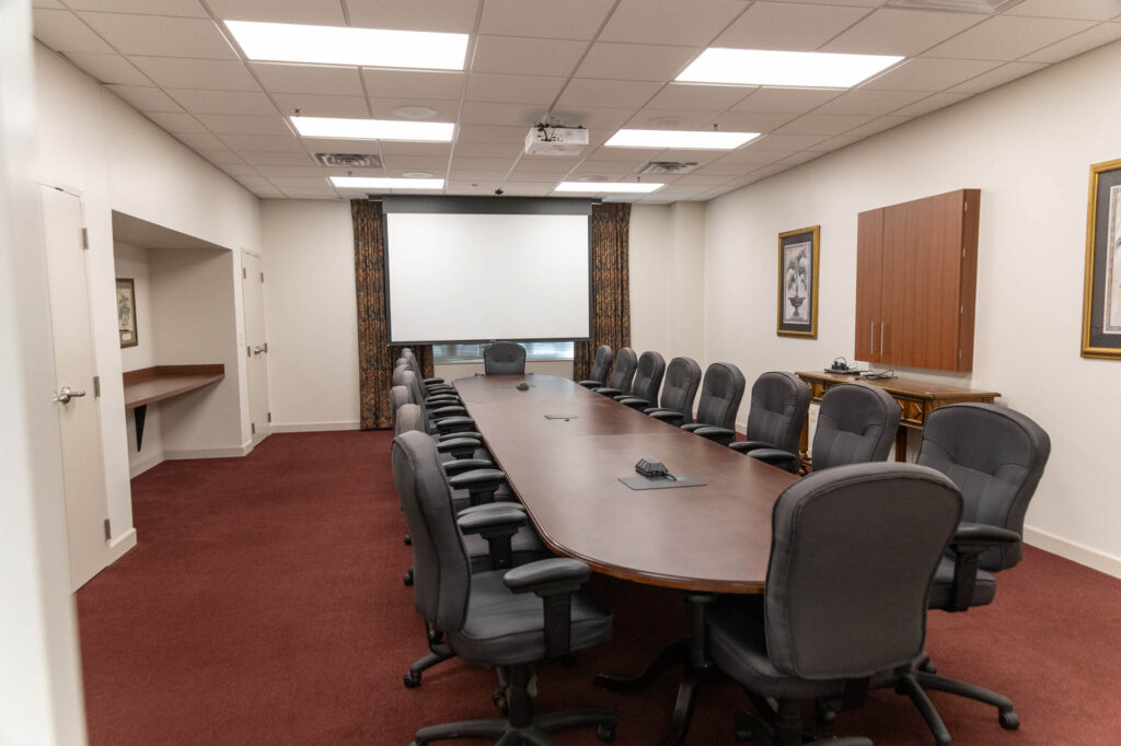A large conference room with a long oval table, surrounded by office chairs. A projector screen is positioned at the far end, with framed pictures and cabinets on the walls. The floor is carpeted in red.