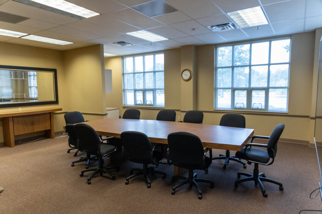 A conference room with a rectangular table surrounded by ten black office chairs, large windows letting in natural light, beige walls, a wall clock, and a mirror on one wall.