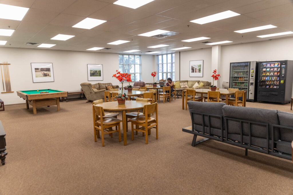 A lounge with tables, chairs, sofas, a pool table, and two vending machines. The room has beige carpet, white walls with framed art, and large windows letting in natural light. One person sits at a table.