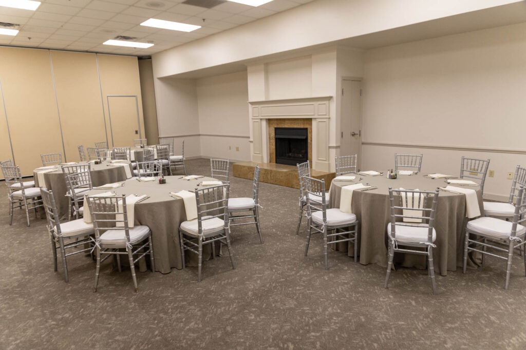 A banquet room with round tables covered in beige tablecloths, each surrounded by silver chairs with white cushions. A fireplace is centered on the far wall. The room is empty and well-lit.