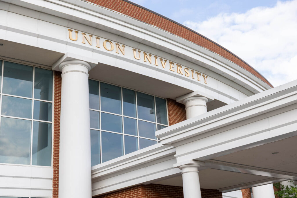 The image shows the entrance of a building with large white columns and windows. Union University is displayed in gold letters above the entrance. The sky is partly cloudy.