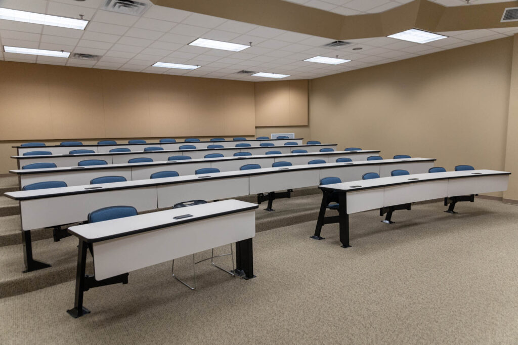 An empty lecture hall with tiered seating, rows of white desks, and blue chairs on a beige carpeted floor. The room has a beige wall, white ceiling tiles, and fluorescent lights.