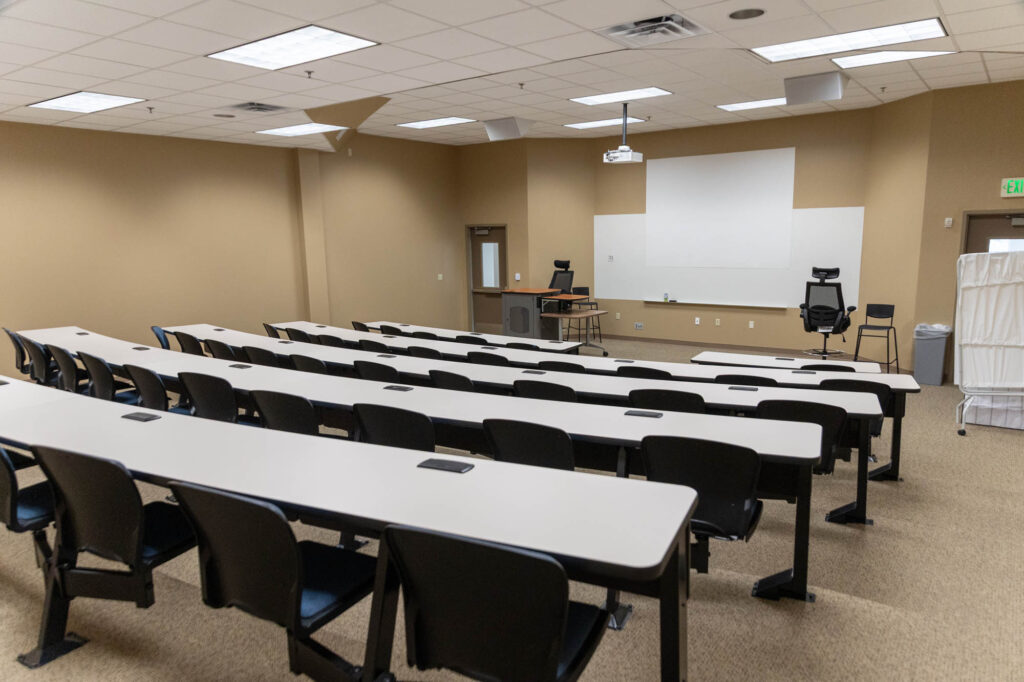 An empty classroom with rows of black chairs and white desks facing a whiteboard, projector, and instructors desk at the front. The walls are beige and the room is well-lit with overhead lights.