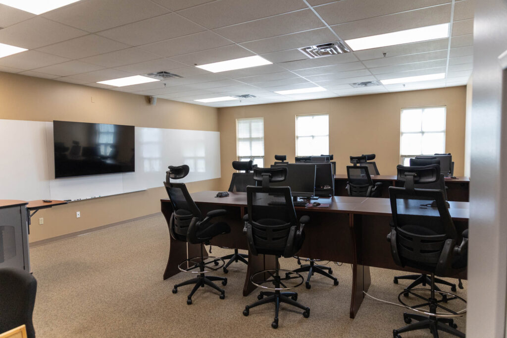 A modern classroom with large desks arranged in a U-shape, each with a computer and office chair. There is a whiteboard and a large wall-mounted screen at the front. The room has beige walls and fluorescent ceiling lights.