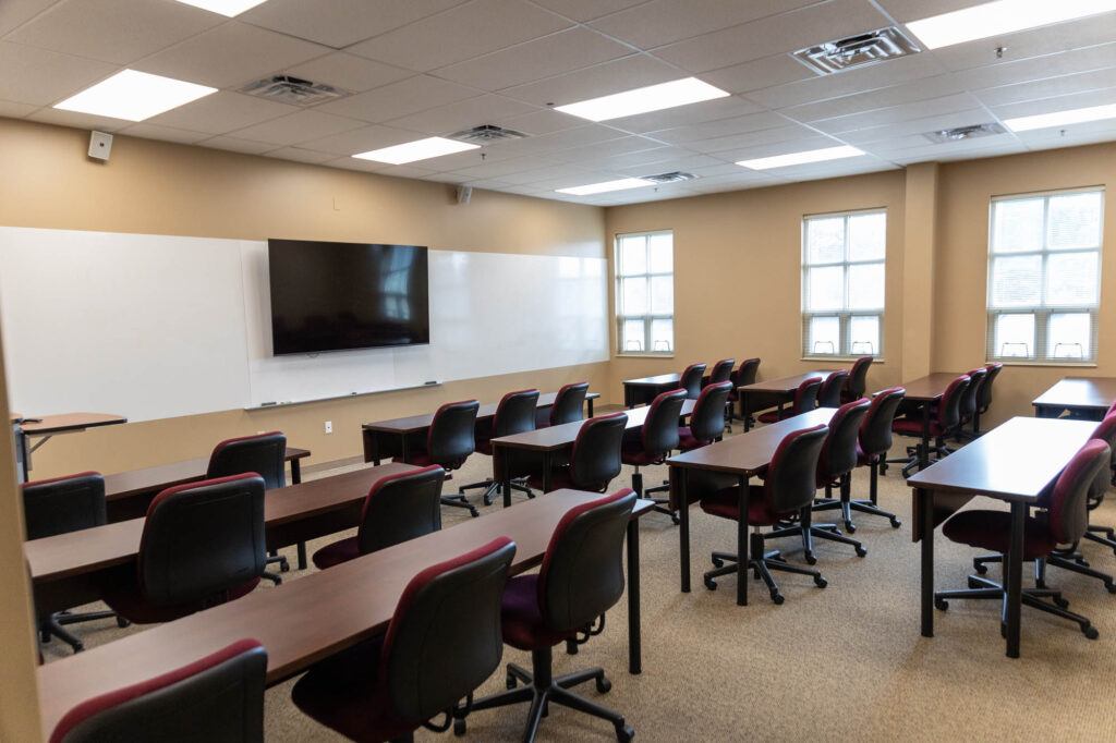 Classroom with rows of desks and chairs facing a whiteboard and a large wall-mounted screen. The room has beige walls, carpeted flooring, and several windows letting in natural light.