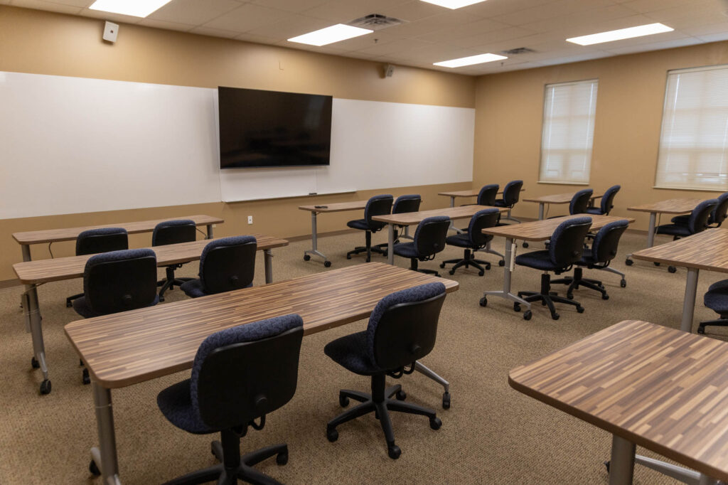 A modern classroom with empty desks and chairs arranged in rows, facing a large wall-mounted screen and whiteboard. The room has beige walls, carpeted floor, and two windows with blinds.