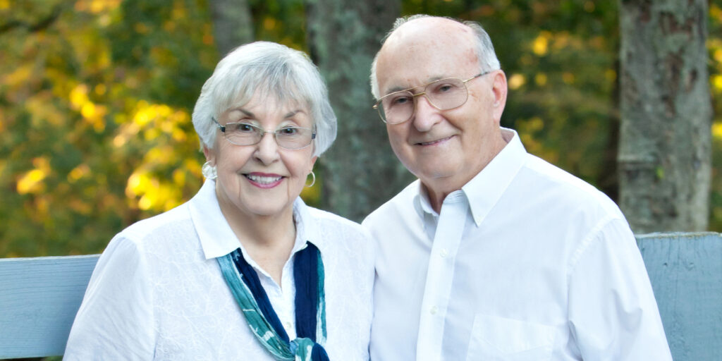 An older woman and man stand outdoors in front of trees, both smiling at the camera. The woman wears glasses, a white shirt, and a scarf; the man also wears glasses and a white shirt.