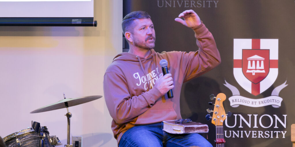 A man in a hoodie speaks into a microphone while seated on stage at Union University during the EQUIP conference. He gestures with his left hand as he encourages youth to speak the truth. Musical instruments and an audience are visible, with a Union University banner in the background.