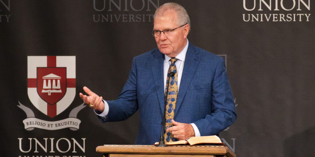 A man in a blue suit and patterned tie stands at a podium with an open book, gesturing as he speaks. The backdrop displays Union University and a red and white shield logo, highlighting the university’s commitment to preparing West Tennessee pastors for impactful ministry.