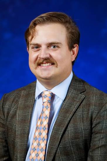 A man with light brown hair and a mustache, wearing a plaid suit jacket, light blue shirt, and a peach and blue checkered tie, poses in front of a plain blue background.