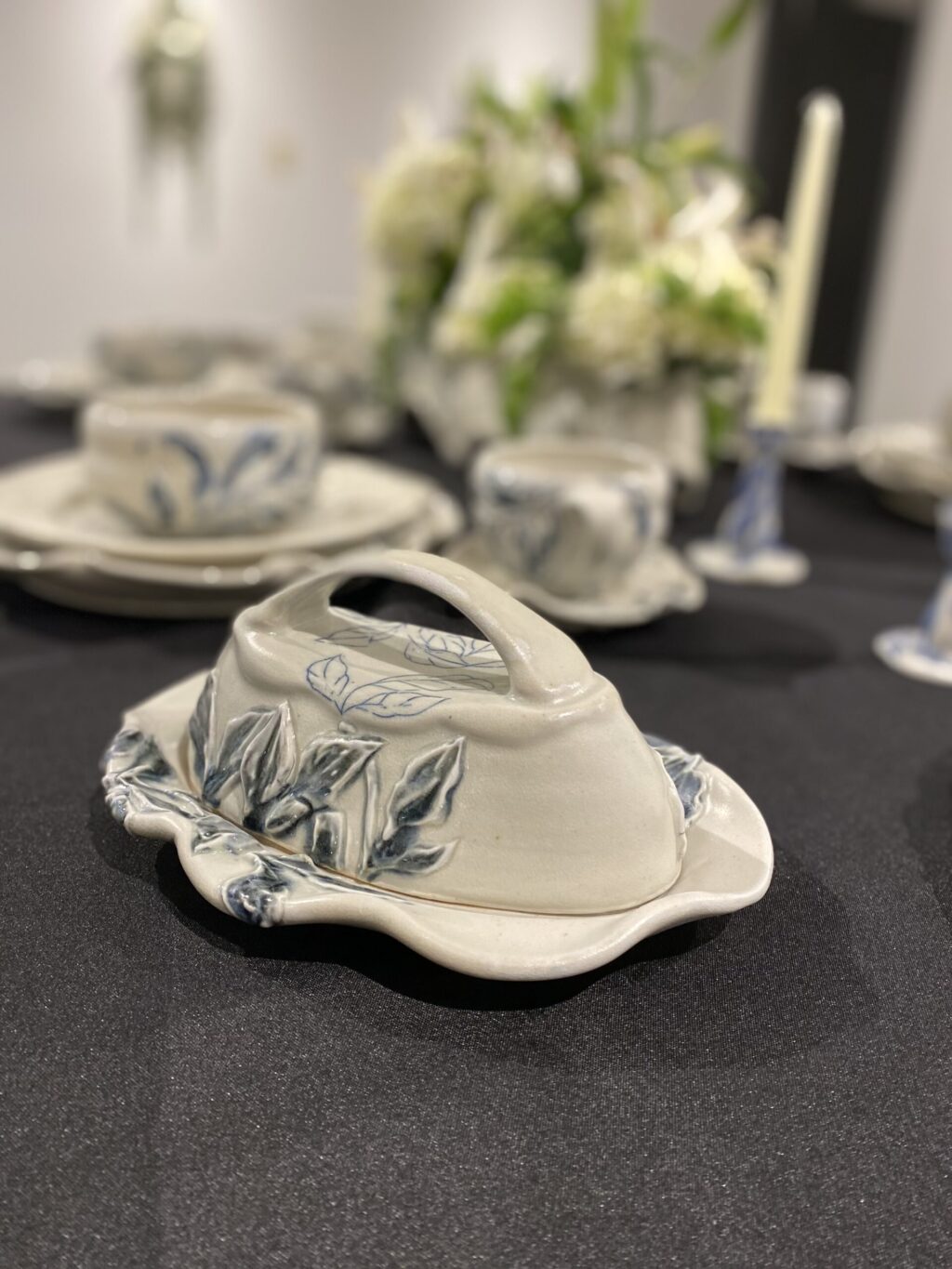 A close-up of a ceramic butter dish with a lid featuring blue leaf designs, placed on a black tablecloth. Blurred matching dishes and floral centerpiece are visible in the background.