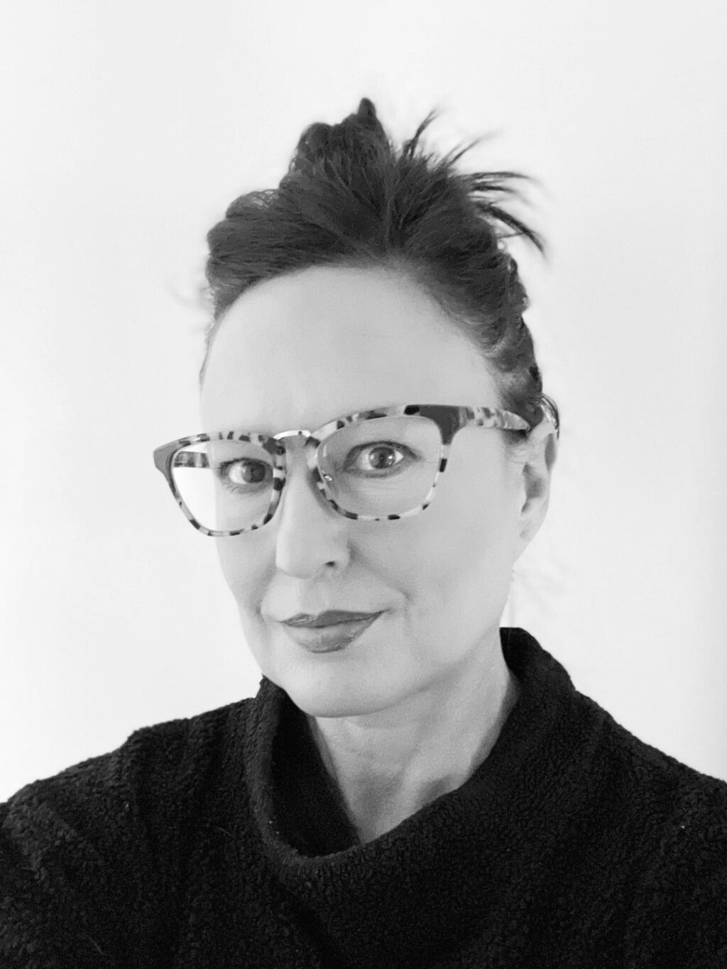 Black and white photo of a person with dark hair in a messy bun, wearing patterned glasses and a textured dark top, looking directly at the camera with a neutral expression against a plain background.