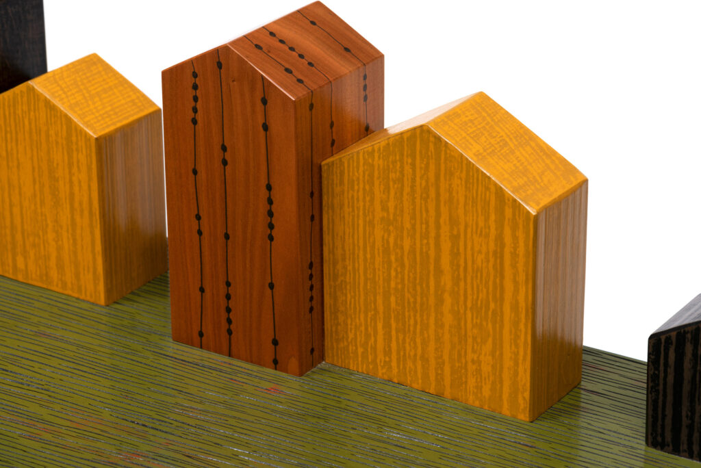 Three wooden blocks shaped like houses, two yellow with roof-like tops and one brown with vertical black dotted lines, are arranged on a green, striped surface against a white background.