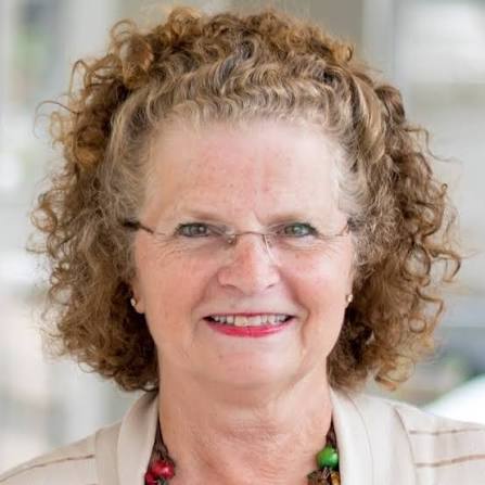 A woman with curly, light brown hair and glasses smiles at the camera. She is wearing a light-colored top and a colorful beaded necklace. The background is softly blurred, suggesting an indoor setting.