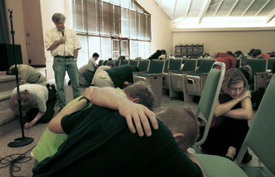 A group of people sit or kneel with heads bowed in a room with rows of chairs, while a man stands at the front near a microphone. Light enters through large windows.