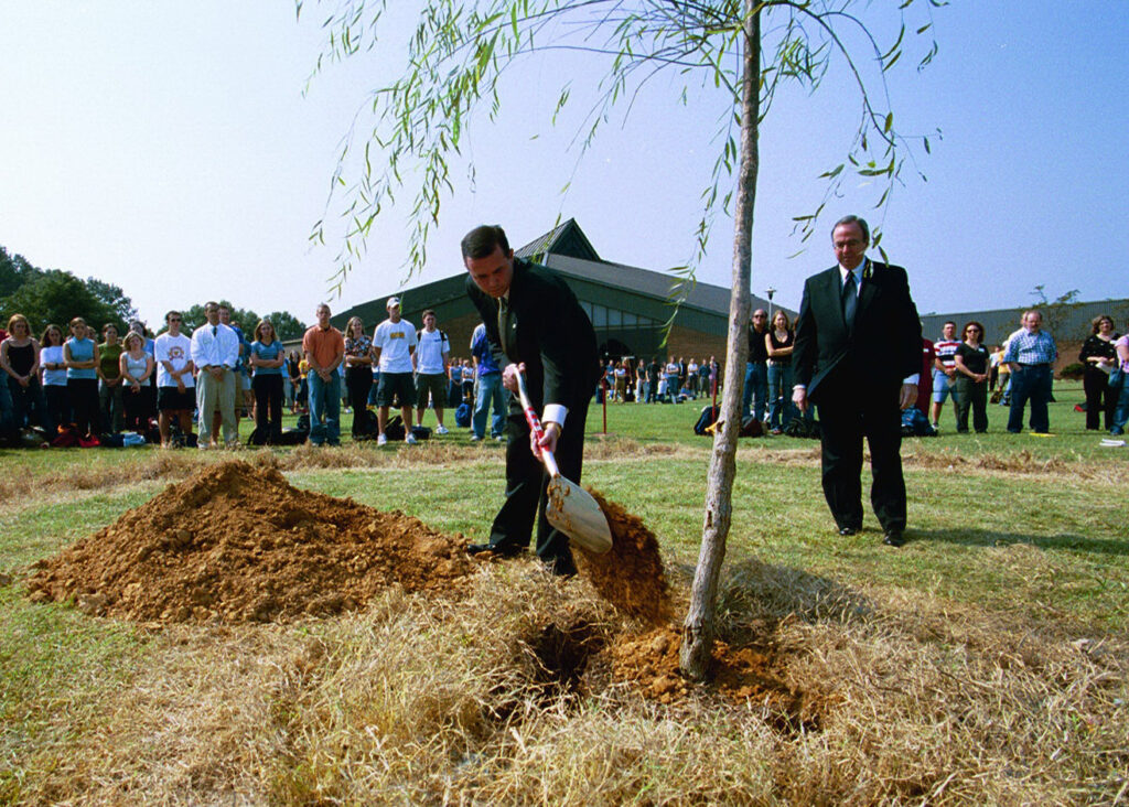 A man in a suit uses a shovel to plant a young tree in a grassy area while another man in a suit stands nearby. A group of people stands in the background, watching the event.