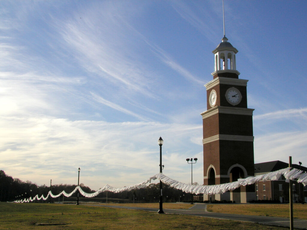 A tall brick clock tower stands on a grassy area under a blue sky. A long string with white fabric pieces or streamers is draped between lampposts leading away from the tower.