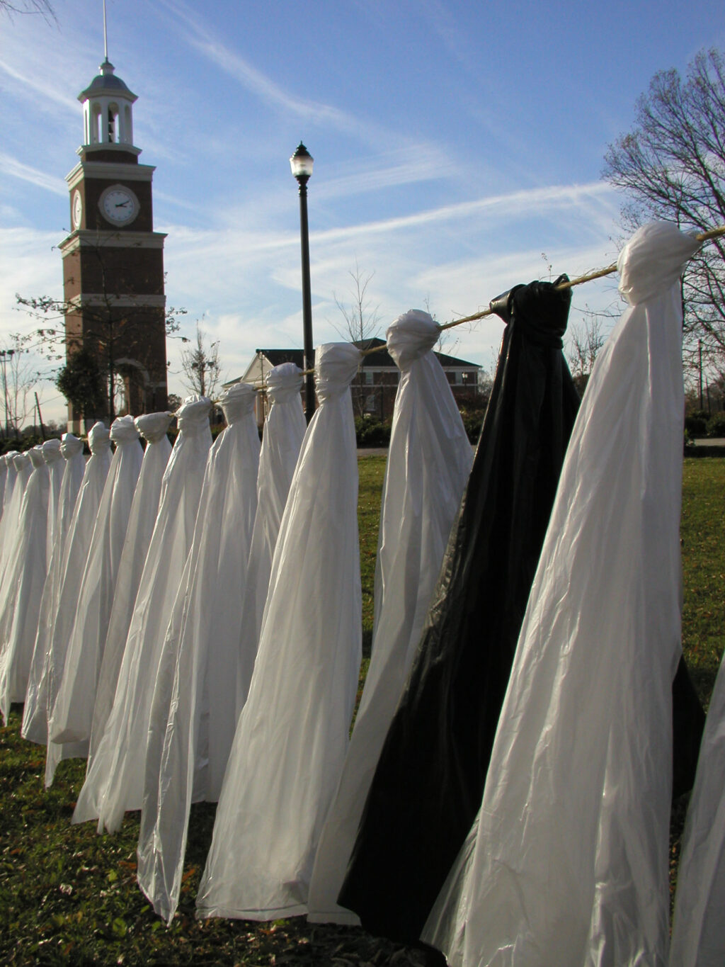 A row of ghost-like figures made from white fabric and one black figure stand on a grassy field, with a clock tower and lamp post visible in the background under a clear sky.