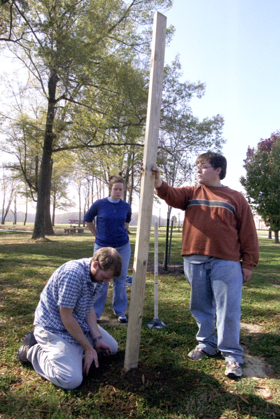 Three people are outdoors on grass by trees. One person kneels to work on the ground, another stands holding a vertical wooden post, and a third person stands in the background watching. A shovel leans against the post.