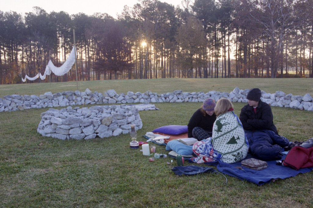 Three people sit on blankets inside a stone circle on a grassy field at sunrise. They are bundled in warm clothing and blankets, with candles and items around them. Trees and hanging fabric are visible in the background.