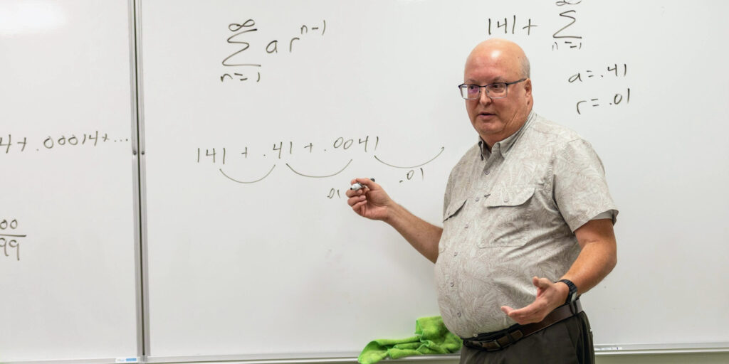 A man stands in front of a whiteboard with mathematical equations written in black marker, holding a marker in one hand and gesturing with the other as if explaining concepts from a calculus textbook to students. A green cloth rests on the tray below the board.