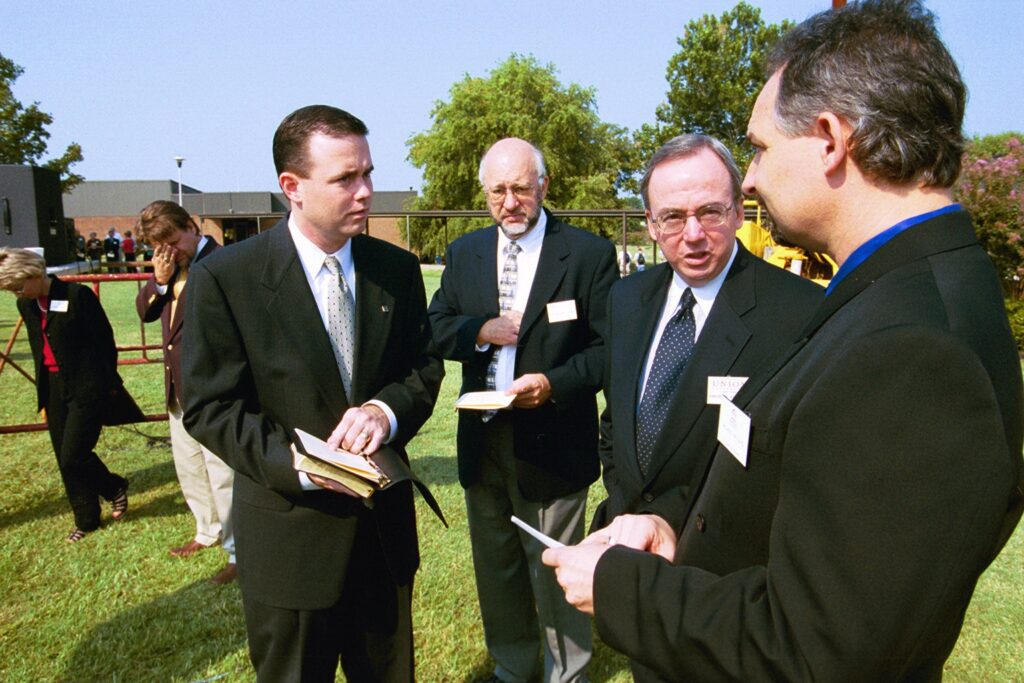 Four men in suits stand on grass having a conversation, holding notebooks and papers. Other people are visible in the background outside near trees and a building on a clear, sunny day.