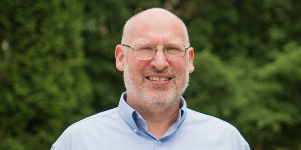 A middle-aged man with glasses, a bald head, and gray beard stands outdoors, smiling. He is wearing a light blue button-down shirt. Green foliage is blurred in the background.