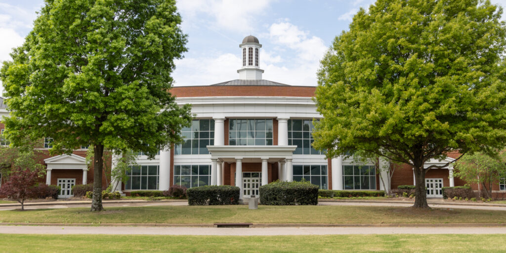 A symmetrical brick building with large windows, white columns, and a cupola on the roof—reminiscent of classic structures found on the Memphis campus Union—is flanked by two tall green trees. The building sits behind a lawn and a sidewalk under a partly cloudy sky.