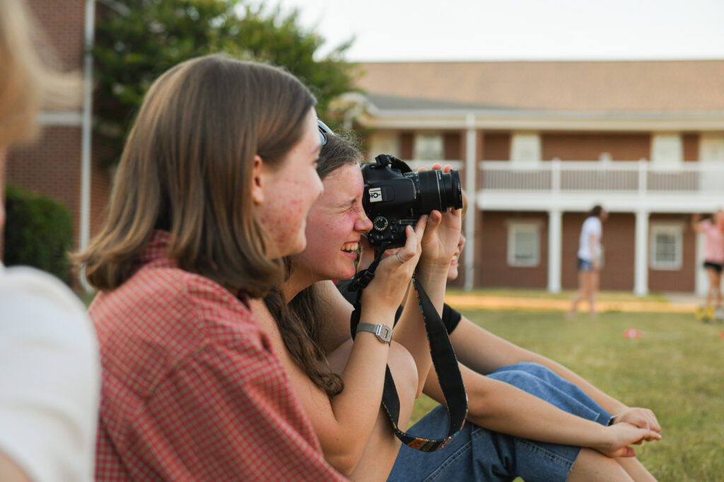 A young person smiles while holding a DSLR camera to their eye, while another person sits beside them.
