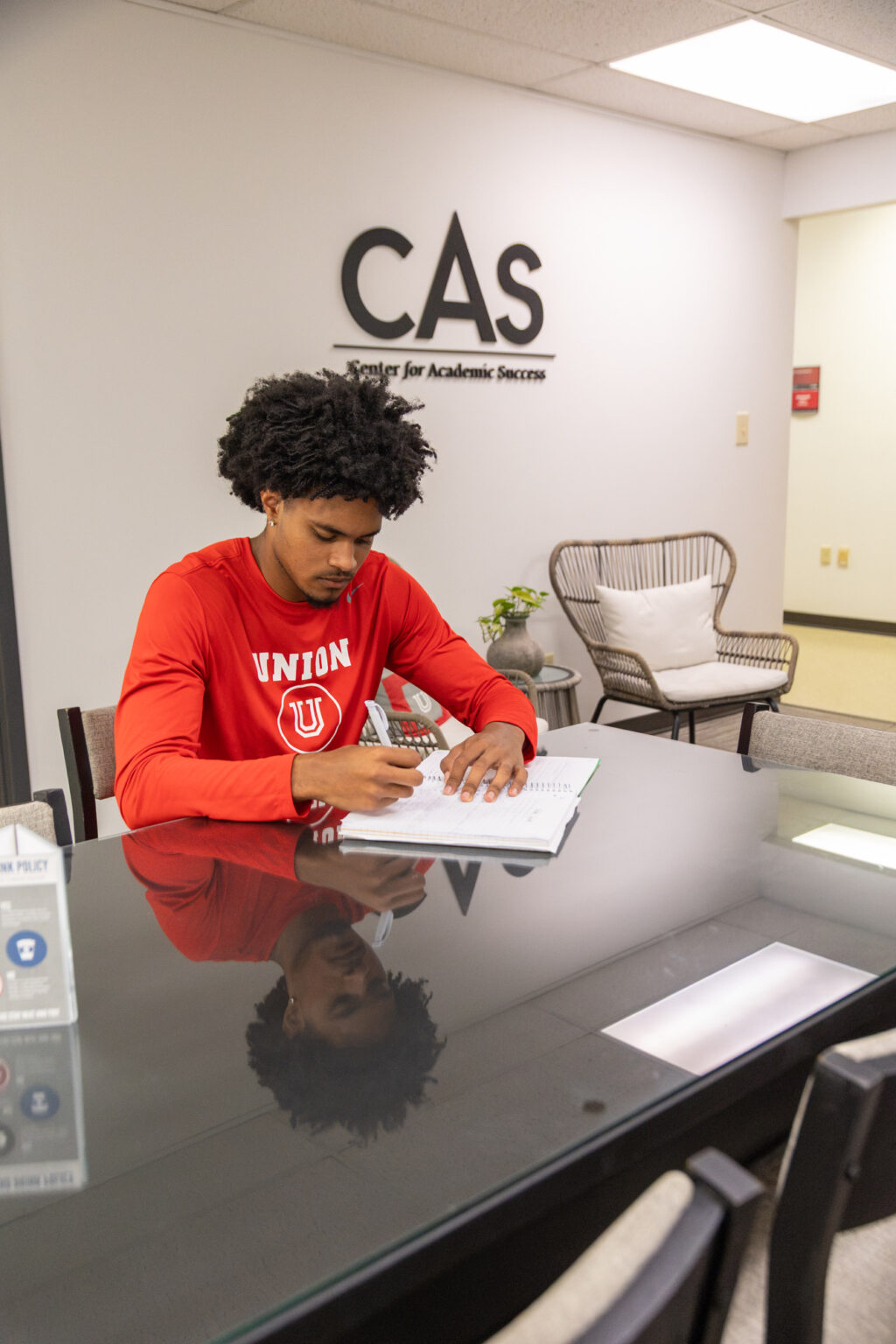A person wearing a red Union University long-sleeve shirt sits at a conference table writing in a notebook in an office labeled Center for Academic Success.
