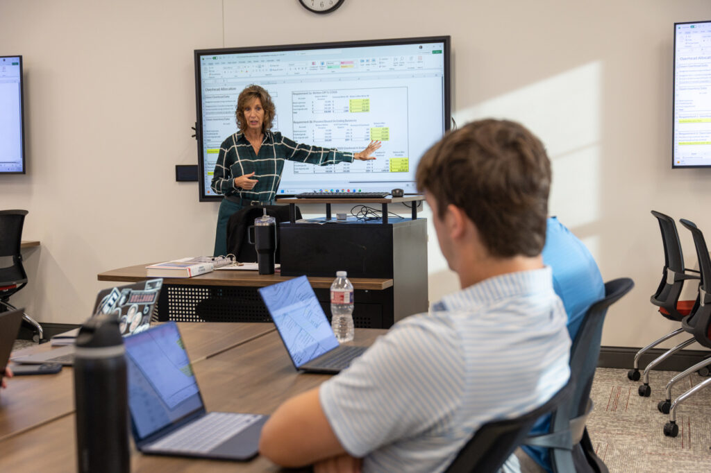 A woman stands at the front of a classroom gesturing to a digital display showing a spreadsheet titled Overhead Allocation while students with laptops listen.