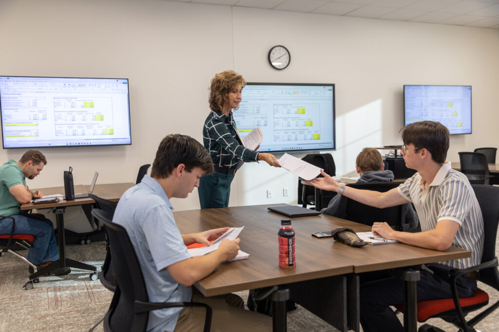 A teacher stands at a conference table in a classroom, handing a document to a student.