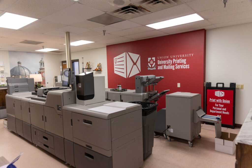 Large commercial printers in a university print shop with a red wall displaying Union University Printing and Mailing Services and a sign for Union Print services nearby. Ceiling lights and some decor are visible.
